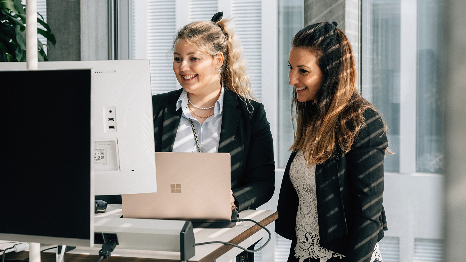 2 Frauen stehen neben einem Tischen und schauen mit einem Lachen auf den Computer.